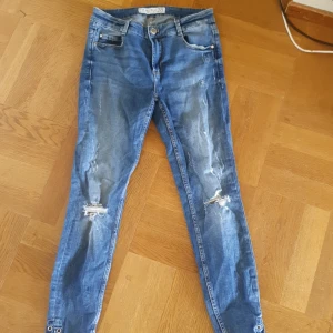 Blå jeansbyxor - Ett par blå jeansbyxor med slitna detaljer på knäna. Byxorna har en knapp och dragkedja framtill samt fem fickor, två fram och två bak.