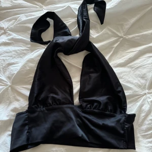 Svart korsett från Gina Tricot - En elegant svart halterbeck från Gina Tricot i storlek XS. Halterneck har en knytning framtill och är tillverkad i ett glansigt material.
