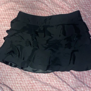 Svart kortkjol med volanger - En svart kortkjol med flera lager av volanger. Kjolen har en dragkedja på sidan och är tillverkad i ett lätt och flödande material.