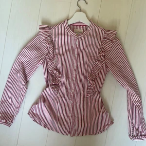 Rosa vit randig blus med volanger - En blus i bra skick, från märket Holly white. Använd fåtal gånger. Den har volanger över axlarna och fina detaljer vid ärmsluten.