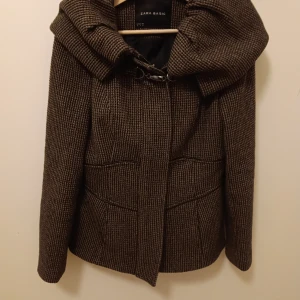 Vårjacka med hög krage - Vårjacka, kragtigt figursydd. Strl 38(M) Dragkedja och knappar fram för snyggast passforn. Hör fluffig krage. Tuget är i robust tyg, typ tweed, i svart/brunt. Zara.