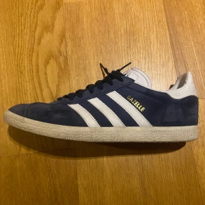 Adidas Gazelle skor 44 - Adidas Gazelle skor 44  Färg: CONAVY/WHITE/GOLDMT Använda fåtal gånger, dom är i utmärkt skick, dom är i storlek 44 men känns lite mindre så dom passar bra på någon med storlek 43. Pris kan diskuteras.
