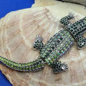 Den här broschen föreställer en krokodil och måtten är ca 14.5 × 4cm. Vikt ca 35g. Silverfärgad metall med vacker ytstruktur och kristaller i flera färger, bl.a grön kulör. Den är ledad i 3 delar. Otroligt vacker brosch av hög kvalitet.  Inkl. Läderh