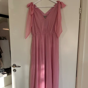 Maxiklänning h&m 40 - Rosa långklänning från H&M i skir, vävd kvalitet med V-ringning fram och bak. Breda axelband med rynk och långa knytband som kan knytas i en rosett upptill. Smal resår i midjan och utställd, böljande kjol. Fodrad. 100% polyester. Storlek 40. Ny.