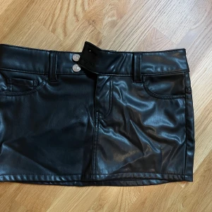 Svart kjol från Divided - En svart kortkjol från Divided i skinnimitation. Kjolen har två framfickor och två bakfickor samt bälteshällor och två knappar i midjan.