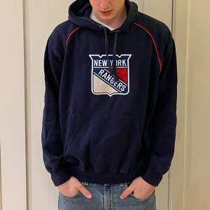 En mörkblå hoodie från Majestic. Storleken är uppskattningsvis medium, M. Hoodien har en logga med New York Rangers, ett amerikanska ishockeylag, på framsidan. Kvaliten är bra.