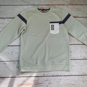 Grön långärmad tröja från Didriksons - Säljer en stilren grön långärmad tröja från Didriksons med en vit bröstficka och mörkgrå detaljer på ärmarna. Perfekt för en avslappnad stil. Tröjan har en rund halsringning och är tillverkad i ett mjukt material. Den har tummhål på slutet av båda ärmarna