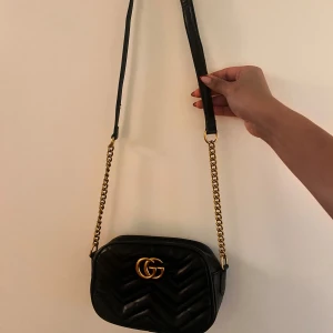 Svart GG  axelväska med gulddetaljer - Snygg svart axelväska i quiltat mönster med guldfärgad kedja och GG-logga. Väskan har en elegant design med hjärtdetalj på baksidan och är perfekt för att lyfta din stil.