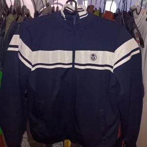 Sergio Tacchini - Snygg navy vindjacka från Sergio Tacchini med vita ränder över bröstet och ärmarna. Jackan har en dragkedja framtill och en hög krage. Perfekt för en sportig look.