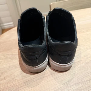 Svarta axel arigato sneakers med dragkedja - Snygga svarta sneakers från Axel Arigato med dragkedja framtill. Skorna har en vit sula och är tillverkade i skinn, vilket ger en stilren look. Perfekta för en avslappnad men trendig stil.