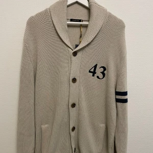 Beige stickad kofta från Batistini - Säljer en stilren beige stickad kofta från Batistini med knappar framtill och två fickor. Den har ett tryck med siffran 43 på bröstet och två mörkblå ränder på ärmen. Perfekt för en avslappnad look.