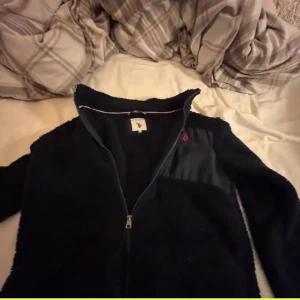 Svart fleecejacka med dragkedja polo Ralph laruren   - Säljer en svart fleecejacka med dragkedja och en liten broderad detalj på bröstet. Jackan har långa ärmar och en hög krage för extra värme. Perfekt för kyliga dagar!