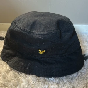 Svart bucket hat från Lyle & Scott - Snygg svart bucket hat från Lyle & Scott med en gul broderad logga framtill. Tillverkad i 100% bomull för en bekväm passform. Perfekt för en stilren look.
