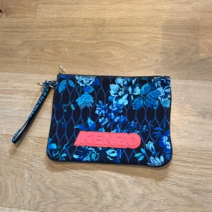 Blommig och leopardmönstrad kuvertväska från Kenzo - Snygg kuvertväska från Kenzo med två olika sidor: en med blått blommönster och en med färgglatt leopardmönster. Väskan har en dragkedja och ett avtagbart handledsband. Perfekt för att ge din outfit en färgklick!