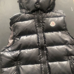 Svart moncler väst - Snygg svart dunväst från Moncler med dragkedja och huva. Perfekt för kyliga dagar när du vill hålla stilen. Västen har Monclers ikoniska logga på bröstet och är både praktisk och trendig.