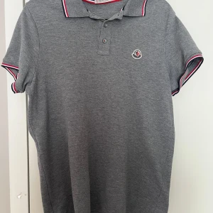 Grå pikétröja med röd och vit detalj - Snygg grå pikétröja från Moncler med klassisk krage och knappar. Tröjan har röda och vita detaljer på krage och ärmslut samt en broderad logga på bröstet. Perfekt för en stilren look!