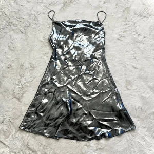 Silverfärgad klänning med smala axelband - Snygg silverfärgad klänning med smala axelband och glansigt material. Perfekt för en kväll ute eller en festlig tillställning. Klänningen har en elegant och modern look.