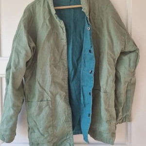 Vändbar linnejacka i grönt och turkos - Säljer en vändbar jacka i 100% linne med en sida i grönt och den andra i turkos. Jackan har knappar framtill och stora fickor på båda sidor. Perfekt för vår och sommar! Märke är Gudrun Sjödén. 
