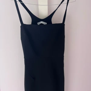 Svart klänning från Zara - Snygg svart klänning från Zara med smala axelband och en stilren design. Klänningen har en öppen rygg och är perfekt för en elegant look. Tillverkad i ett mjukt och bekvämt material.