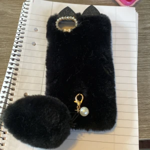 Svart fluffigt mobilskal med pärldetalj - Säljer ett svart fluffigt mobilskal med öron och en pärldetalj. Skalet har en glittrig kant runt kameran och en fluffig boll som hänger nedtill. Perfekt för att ge din mobil en unik och mysig look!