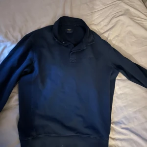 Mörkblå tröja från Superdry - Säljer en mörkblå tröja från Superdry med en stilren design. Tröjan har långa ärmar och en hög krage med knappdetalj. Perfekt för en avslappnad stil.