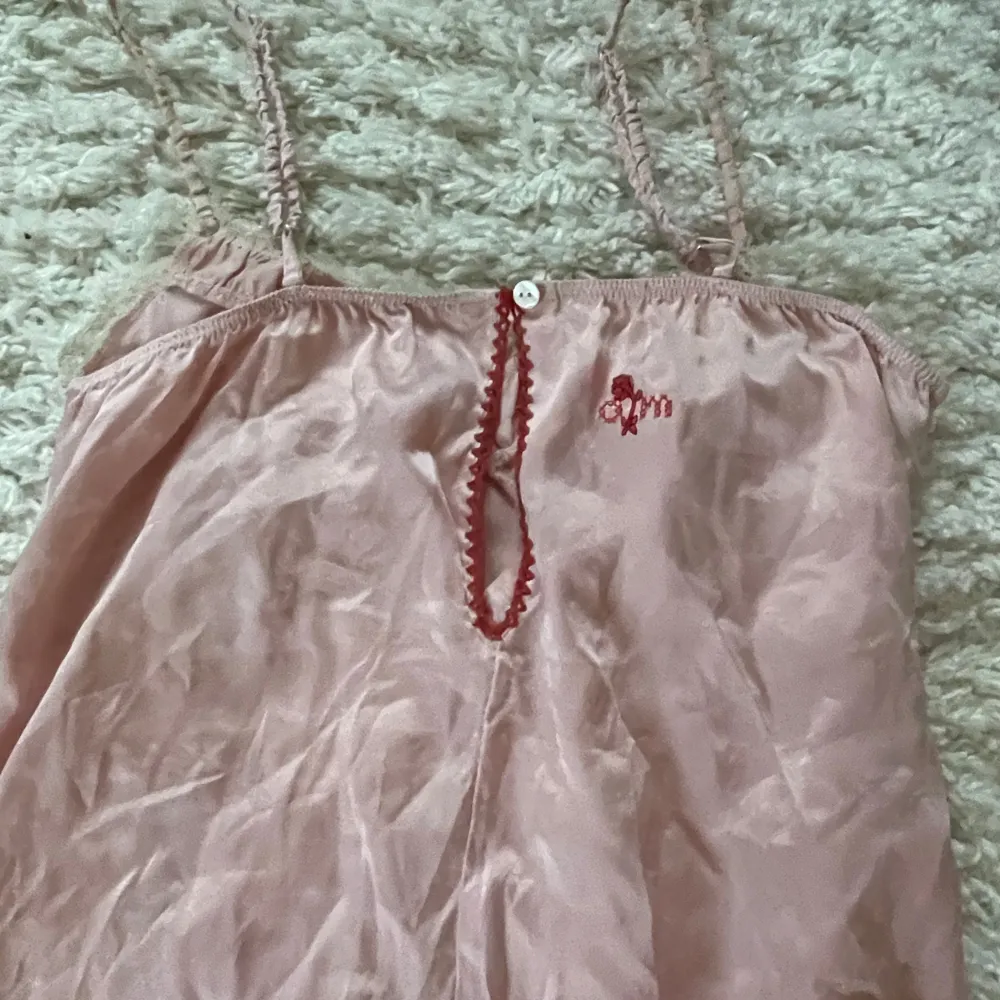 Elegant rosa klänning/nattlinne i siden med smala axelband och spetsdetaljer vid fållen. Klänningen har en liten rosett framtill och en dekorativ öppning med knappdetalj på baksidan.. Mekot.