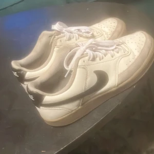 Vita Nike court vison - Säljer ett par klassiska vita Nike sneakers med svart logo och kaffe brun sula nu de är för små. Skorna har en ny snörning och en rund tå.  Priset är ej hugget i sten