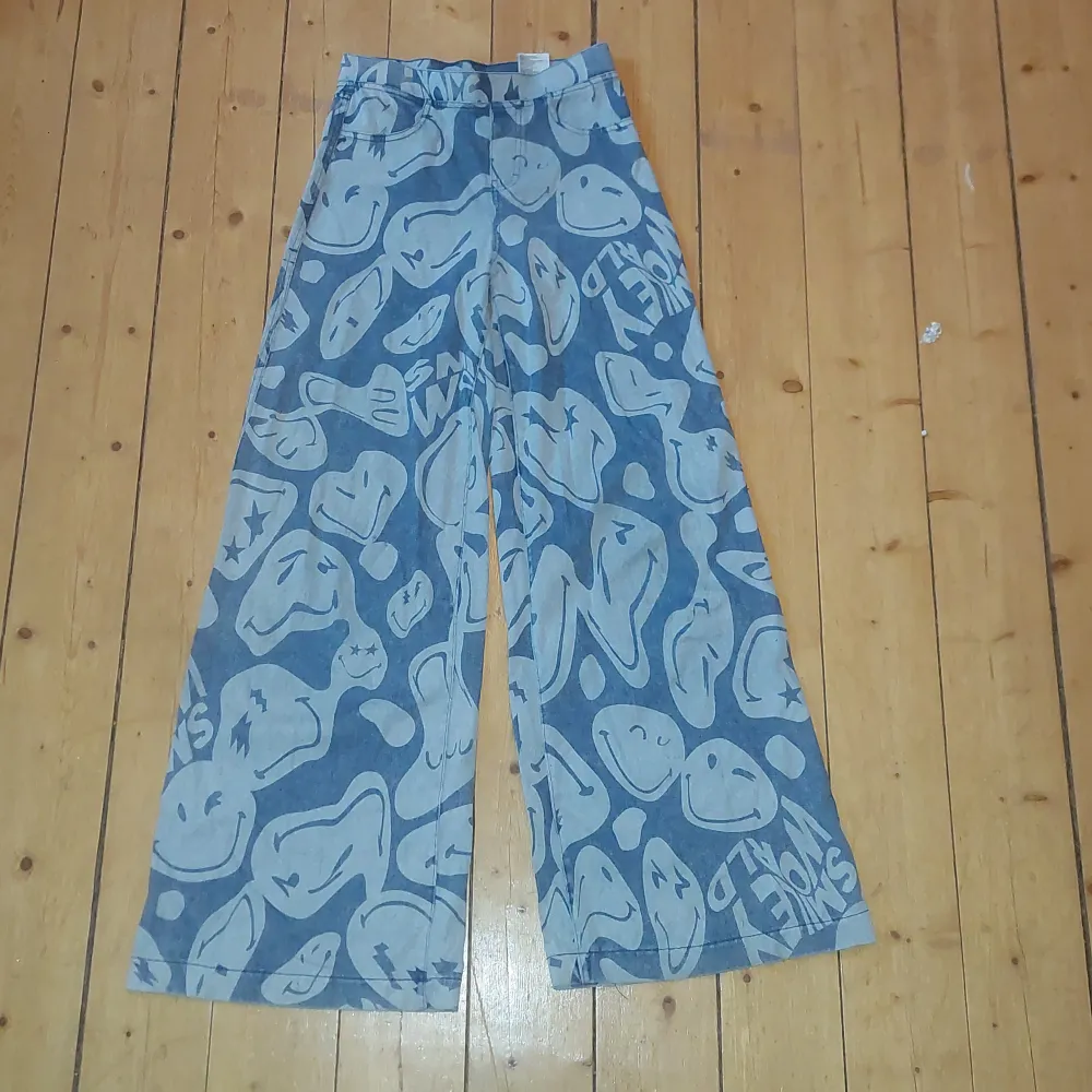 Coola blå byxor från H&M med ett unikt smiley-mönster över hela tyget. Byxorna har en loose passform och är perfekta för en avslappnad stil. De har elastisk midja för extra komfort.. Farkut & Housut.