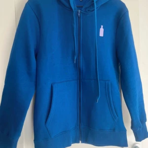 Blå hoodie från Absolut - Säljer en blå hoodie från Absolut med dragkedja och en liten logga på bröstet. Huvan har texten 'ABSOLUT' tryckt på baksidan. Tillverkad i en bomulls- och polyesterblandning för en bekväm passform. Perfekt för en avslappnad stil.