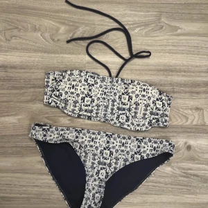 Mönstrad bikini i blått och vitt 38 - Snygg bikini med ett unikt mönster i blått och vitt. Överdelen är bandeau-stil med knytband runt nacken och knäppning i ryggen. Underdelen är klassisk med samma mönster. Perfekt för en dag på stranden!