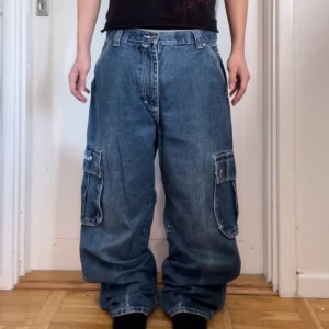 Blå baggy jeans med cargofickor från PJ Mark - Säljer ett par blå baggy jeans från PJ Mark med stora cargofickor på sidorna. Byxorna har en avslappnad passform och är perfekta för en casual look. Broderad logga på baksidan ger en extra touch.