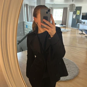 Svart trenchcoat med knappar - Snygg svart trenchcoat från Vero Moda med dubbelknäppning och klassisk krage. Jackan har en stilren design med knappar och fickor framtill. Perfekt för en elegant look.