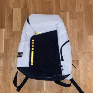 Vit och svart ryggsäck från Nike - Snygg ryggsäck från Nike i vit och svart med guldiga detaljer. Ryggsäcken har flera fack och justerbara axelremmar. Perfekt för både träning och vardag. Märket 'ELITE' syns tydligt på baksidan.