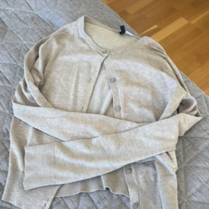 Beige kofta med knappar från H&M - Säljer en stilren beige kofta från H&M med rund hals och knappar framtill. Koftan har långa ärmar och är gjord i mjuk bomullsmix som känns skön mot huden. Perfekt att slänga på sig när det är lite kyligt ute.