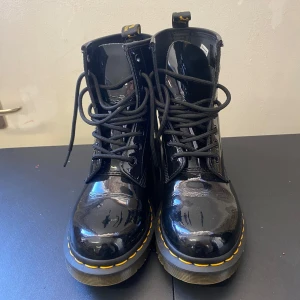 Svarta Dr. Martens 1460, lack - Säljer ett par svarta Dr. Martens 1460 kängor i lackat skinn med klassisk gul söm runt sulan. Modellen har rund tå, snörning och grov platt sula. Perfekt för dig som vill ha en edgy och cool stil med extra glans. Storlek EU 36.
