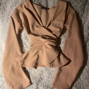 Beige/rosa omlottopp med volang från Boohoo - Supersnygg beige omlottopp från Boohoo med långa ärmar och volang vid midjan. Toppen har knyt i ryggen och slits vid ärmsluten för extra vibe. Materialet är mjukt och har en lätt struktur som ger en cool look.