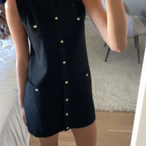 Svart ärmlös kortklänning med knappar - Snygg svart ärmlös kortklänning med raka linjer och guldiga knappar framtill. Super nice nu till hösten. Helt oanvänd