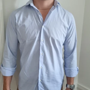 Ljusblå klassisk skjorta i bomull - Stilren ljusblå skjorta med klassisk krage och knappar framtill. Skjortan är långärmad och designad av danska märket Selected Homme. Passformen är normal och skjortan funkar perfekt till jeans, chinos eller kostym.