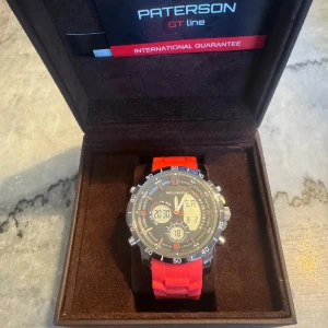 Paterson GT Line klocka med rött armband - Oanvänd Sportig Paterson GT Line klocka med digital och analog urtavla. Svart boett med detaljer i silver och rött, samt ett brett rött gummiarmband. Klockan har flera digitala displayer och tydliga siffror, perfekt för dig som gillar en modern och tuff stil.