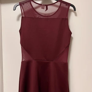 Vinröd ärmlös klänning med meshdetaljer - Snygg vinröd klänning med ärmlös design och transparent mesh upptill och vid midjan. Klänningen har en rund halsringning och en stilren, figurnära passform som ger en modern look. Perfekt för dig som gillar minimalistisk stil med en twist.