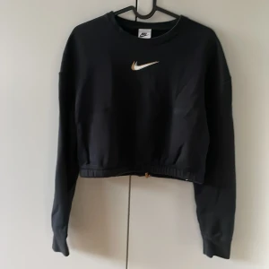 Svart croppad Nike sweatshirt XS - Svart croppad sweatshirt från Nike med loose fit och snyggt broderad logga i vitt och guld på bröstet, samt liten logga på ryggen. Tröjan har långa ärmar och snörning i nederkant för att justera passformen. Perfekt för chill eller träning.