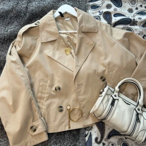Beige croppad trenchcoat från H&M - Snygg croppad trenchcoat i beige från H&M med dubbelknäppning, bred krage och axelklaffar. Jackan har långa ärmar, två fickor fram och coola knappar i mörkare ton. Perfekt för dig som gillar klassisk stil med modern twist.