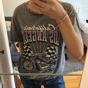 Grå t-shirt  - Cool grå t-shirt från FB Sister med stentvättad look och stort tryck framtill med texten 'California Los Angeles', lila detaljer och en motorcykel med svartvita flaggor. Modellen är kortärmad och har en baggy passform. Perfekt   som streetstyle och statement-plagg💫