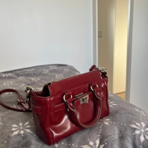 Vinröd axelväska med silverdetaljer - Snygg vinröd axelväska i skinn med både handtag och justerbar axelrem. Väskan har ett stilrent lås framtill, silverfärgade metalldetaljer och dragkedja upptill. 