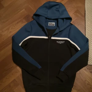 McKenzie zip hoodie - Säljer en snygg hoodie från McKenzie i svart och mörkblått med vit detalj över bröstet. Tröjan har huva, dragkedja och långa ärmar. Perfekt för chill dagar eller när du vill ha en sportig look. Materialet är mjukt och känns skönt mot huden.