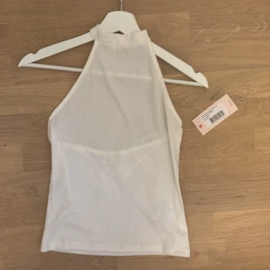 Vit high neck topp från Nelly - Säljer en vit high neck topp från Nelly i storlek small. Toppen är ärmlös med halterneck och knäpps med tryckknapp i nacken. Materialet är strukturerat och ger en cool textur. Perfekt för dig som gillar minimalistisk och trendig stil.