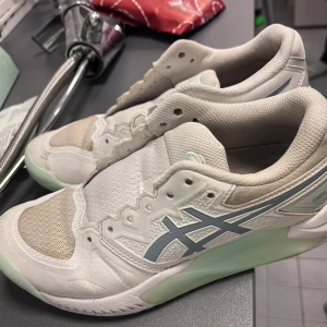 Vita Asics sneakers med mesh och snörning - Fräscha vita Asics sneakers med ljusblå detaljer och logga på sidan. Skorna har ventilerande meshpartier, snörning och en mjuk, dämpad sula i syntet. Perfekta för dig som gillar sportig stil och komfort. Snygg design med rund tå och platt sula. KOMMER MED TVÅ SNÖREN! Köptes för 900kr