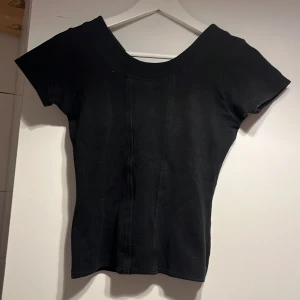 Svart topp från Jeffrey Rogers S - En svart, figurnära topp från Jeffrey Rogers i storlek S. Toppen har korta ärmar, v-ringning i ryggen och bredare rundad halsringning fram. Snygga sömdetaljer framtill ger en cool look. Perfekt att styla med jeans eller kjol.