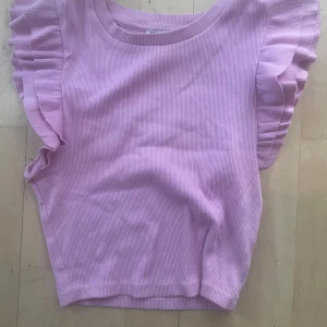 Ljusrosa ribbad topp med volangärm - Supersöt ljusrosa ribbad topp från Zara med breda volangärmar som ger en extra touch. Toppen är ärmlös med rund halsringning och har en mjuk, stretchig känsla. Perfekt för dig som gillar en feminin och trendig stil.