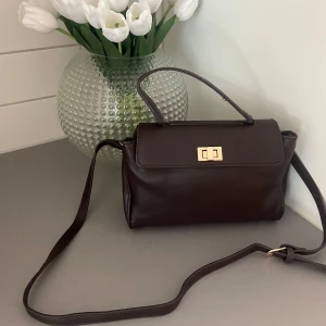 Mörkbrun axelväska med gulddetaljer - OANVÄND!!! 🌟Snygg mörkbrun axelväska i skinn med stilren, rektangulär form och guldfärgat lås framtill. Väskan har både handtag och justerbar axelrem, samt flera fack inuti med dragkedja. 